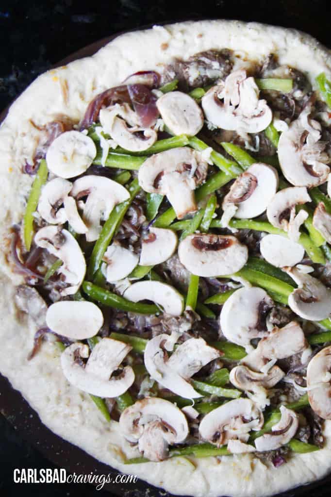 showing how to make Philly Cheesesteak Pizza by layering pizza with bell peppers and mushrooms