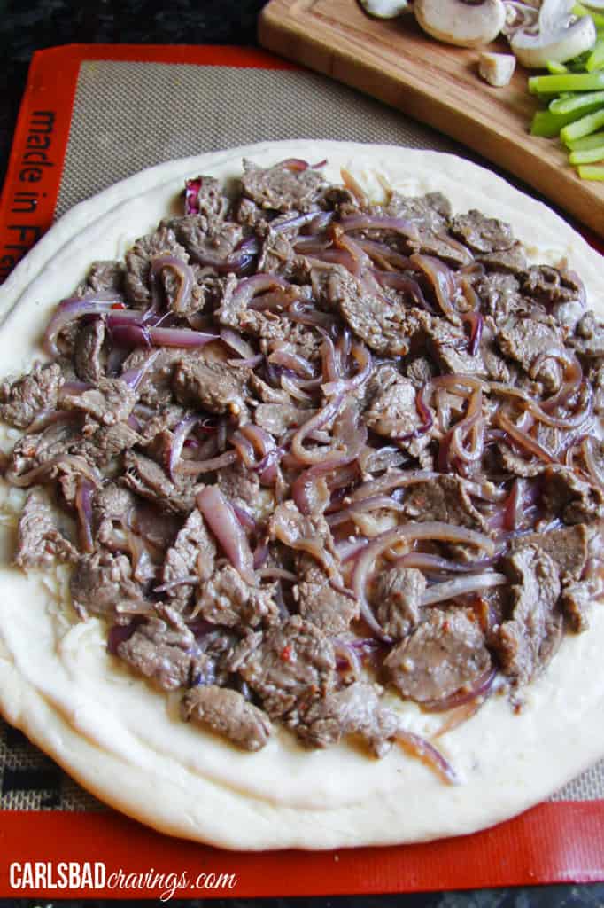 showing how to make Philly Cheesesteak Pizza by layering Alfredo sauce with cheese, steak and onions