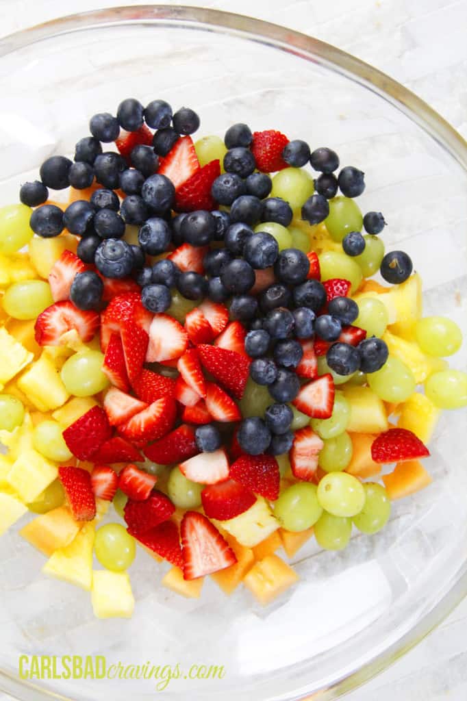 showing how to make fruit salad with yogurt by adding pineapple, grapes, blueberries, strawberries to a bowl
