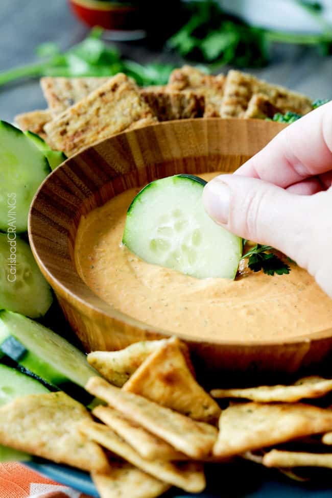 A cucumber being dipped in a bowl of Roasted Red Pepper Feta Dip.