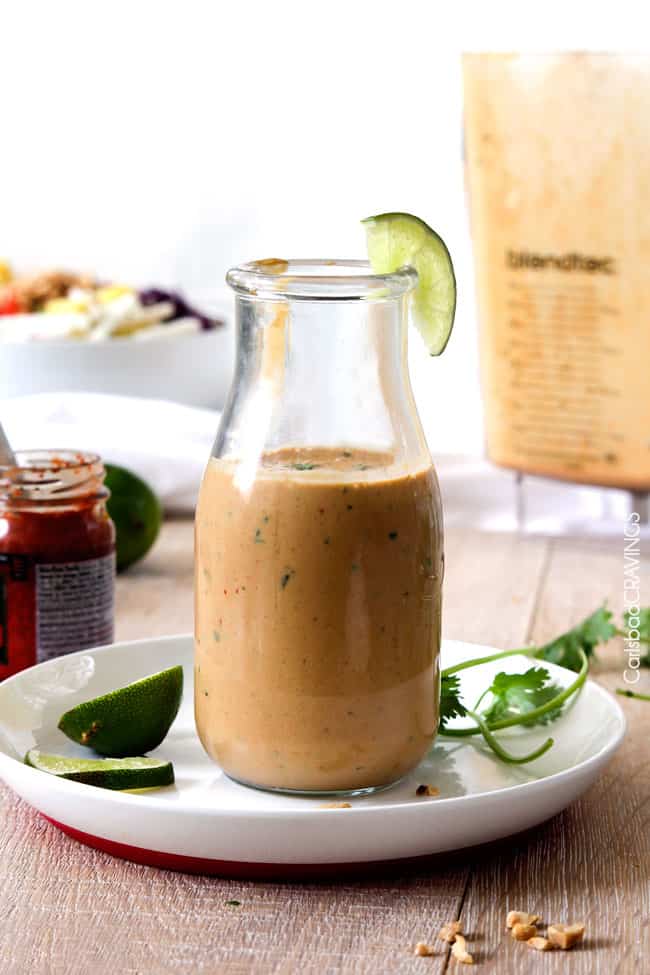 up front view of spicy thai peanut dressing recipe in a glass bottle garnished with limes and cilantro on a white plate