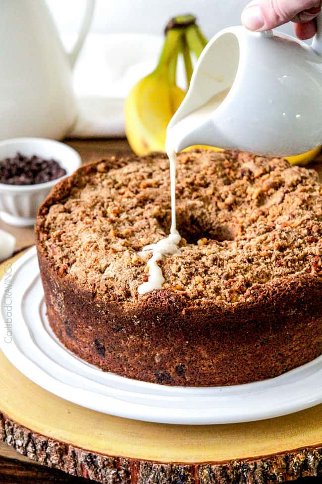 pouring vanilla glaze over best coffee cake on a whiite platter The best Banana ANYTHING ever! Moist Banana Coffee Cake riddled with chocolate chips and walnuts (optional) with an INCREDIBLY creamy cheesecake-like cream cheese filling all topped with brown sugar walnut streusel and vanilla drizzle.