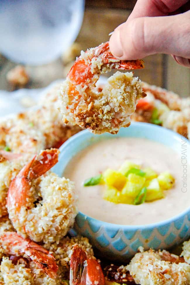 Baked crunchy, juicy Cashew Coconut Shrimp with Pineapple Sweet Chili Dip way better than takeout without the extra fat or cost! Holding a large Coconut Shrimp over a platter next to dipping sauce.