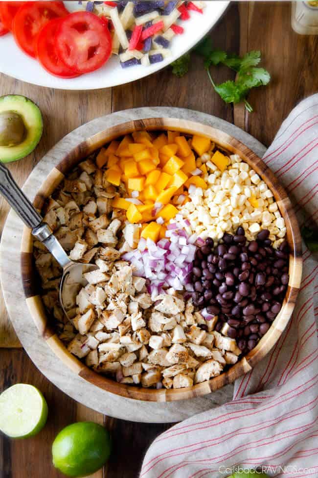 These Avocado Chicken Salad Sandwiches are my favorite way to use leftover chicken! They are super easy, creamy, flavorful, refreshing and I love the Mexican spices and crunch of tortilla strips! Showing how to make Avocado Chicken Salad by adding filling ingredients of chicken, mangos, corn, black beans, to a large wood bowl