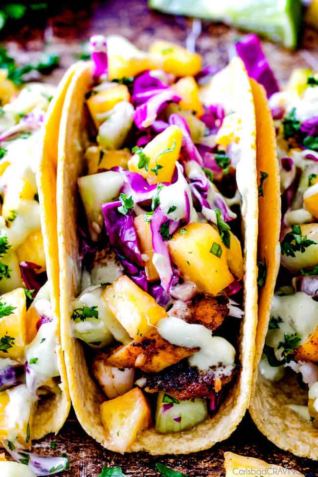 Mind blowing Blackened Tilapia Fish Tacos with a quick marinade and the most flavorful spice rub! These are hands down the BEST FISH TACOS ever! And the sweet and tangy Pineapple Cucumber Slaw and Avocado Crema hit this one out of the park! seriously better than my favorite restaurant! up close front view of easy Tilapia Fish Tacos with cabbage, avocados and cilantro