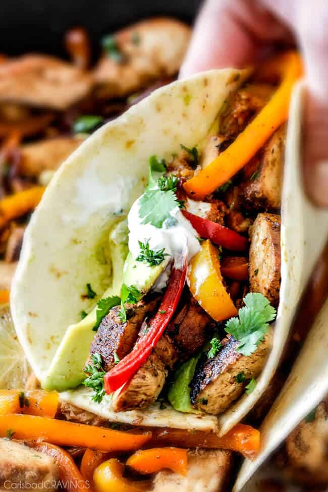 easy Skillet Chicken Fajitas - these are the BEST chicken fajitas! the marinade is seriously the best I've ever tried - better than any restaurant and only 10 minute cook time! My family LOVES this filling so much we use it in quesadillas, burritos, and salads! a hand holding a chicken fajita with sliced chicken, bell peppers, onions, avocados, cilantro and sour cream