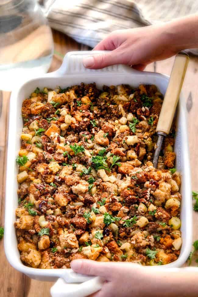 Thanksgiving Cornbread Sausage Stuffing (Dressing) with dried cranberries, apples and pecans is is destined to become your new go-to recipe!! The wonderfully savory, buttery herb infused stuffing is moist, flavorful, easy and absolutely addicting! two hand holding a white casserole dish of the best Cornbread Stuffing with sausage
