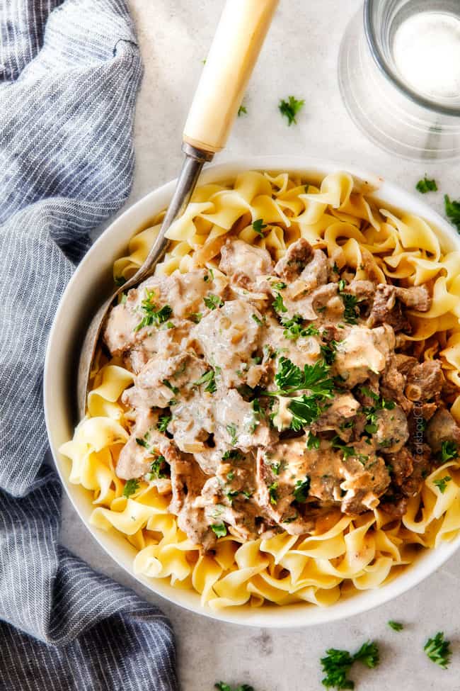 This is by far my family's favorite Slow Cooker Beef Stroganoff recipe! An ultra rich and creamy, amazingly flavorful sauce (without any "cream of" anything!), crazy tender meat all made in the crockpot for a gourmet meal with hardly any effort! I love serving this for holidays like Christmas! top view of homemade Crock Pot Beef Stroganoff on egg noodles in a white bowl with a silver servings spoon and blue napkin