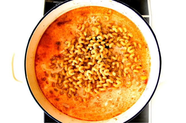 Lighter ONE POT Chicken Fajita Pastaย has only minutes of hands-on cooking time! ย Its smothered in cheesy, creamy salsa infused sauce bursting with your favorite fajita spiced chicken, onions and peppers! ย The pasta cooks right IN the sauce for simple prep, minimal cleanup, and extra flavooooor! ย showing how to make healthy Chicken Fajita Pasta by adding pasta to pot with milk, salsa and enchilada sauce.