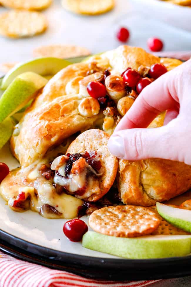 Rich and creamy Baked Brie in Puff Pastry is always a total luxuriously tasting crowd pleaser with minimal effort!ย (step by step pictures included)ย It can be prepped in advance then baked when you're ready for company!ย scooping a bite of baked brie in puff pastry with cranberries with a cracker