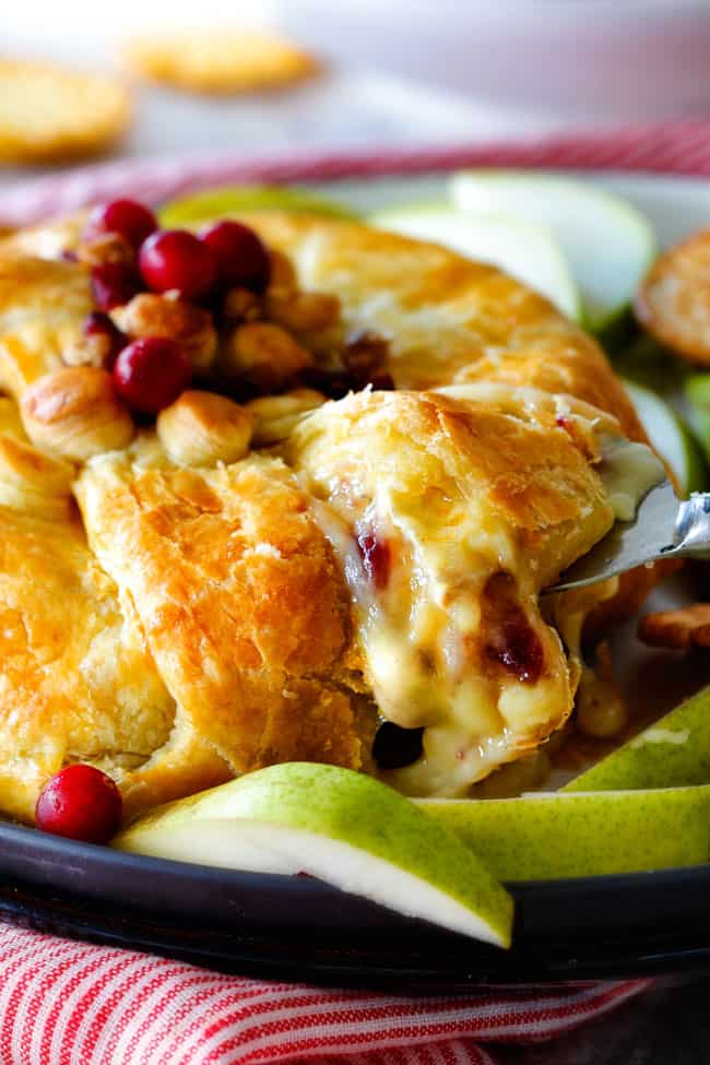Rich and creamy Baked Brie in Puff Pastry is always a total luxuriously tasting crowd pleaser with minimal effort!ย (step by step pictures included)ย It can be prepped in advance then baked when you're ready for company!ย a serving spoon taking a slice of baked brie in puff pastry with cranberries and jam