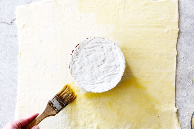 Rich and creamy Baked Brie in Puff Pastry is always a total luxuriously tasting crowd pleaser with minimal effort!ย (step by step pictures included)ย It can be prepped in advance then baked when you're ready for company!ย aaaaaaaaa showing how to make baked brie with puff pastry by laying puff pastry on a flat surface with brie wheel in the center