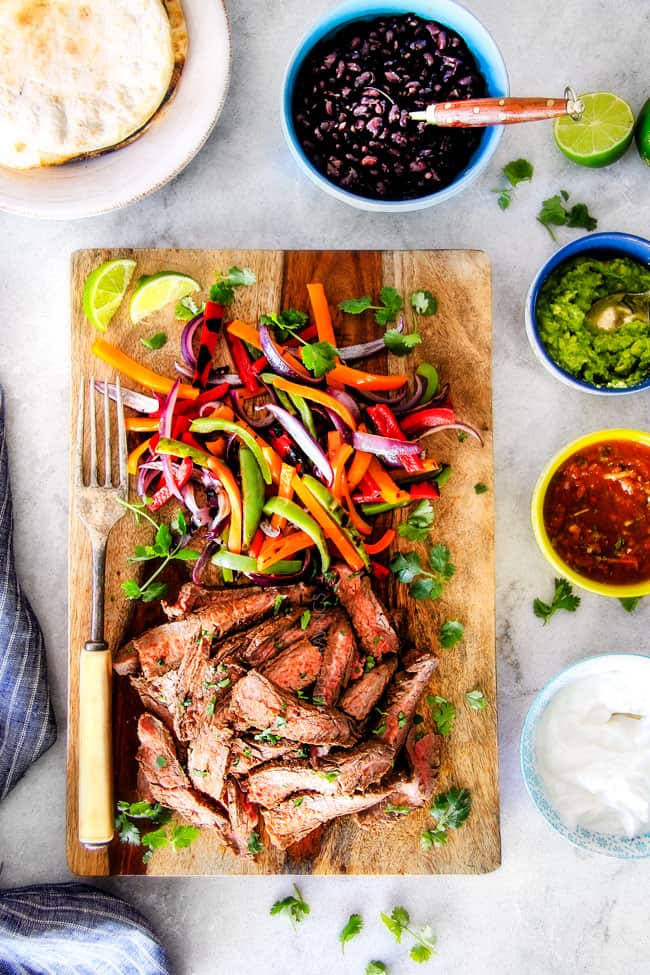 showing how to make steak fajitas by assembling sliced steak and bell peppers on a wood cutting board surrounded by toppings: cilantro, guacamole, salsa and sour cream