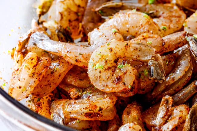 Cilantro Lime Shrimp Tacos EXPLODING with tangy, punchy flavor complimented by sweet refreshing Mango Salsa and silky Avocado Crema on your table in 35 minutes!ย You can prep the toppings ahead of time for a super quick and easy weeknight dinner that tastes better than any restaurant! showing how to make Shrimp Tacos by tossing raw shrimp with lime zest, garlic and seasonings in a glass bowl