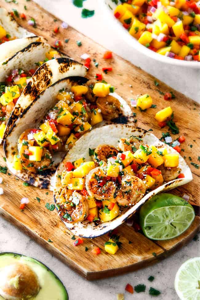 Cilantro Lime Shrimp Tacos EXPLODING with tangy, punchy flavor complimented by sweet refreshing Mango Salsa and silky Avocado Crema on your table in 35 minutes!ย You can prep the toppings ahead of time for a super quick and easy weeknight dinner that tastes better than any restaurant! best Shrimp Tacos with Mango Salsa and Avocado lined in a row on a brown cutting board