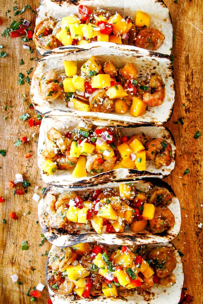 Cilantro Lime Shrimp Tacos EXPLODING with tangy, punchy flavor complimented by sweet refreshing Mango Salsa and silky Avocado Crema on your table in 35 minutes!ย You can prep the toppings ahead of time for a super quick and easy weeknight dinner that tastes better than any restaurant! top view of easy shrimp tacos recipe lined in a row on a brown cutting board