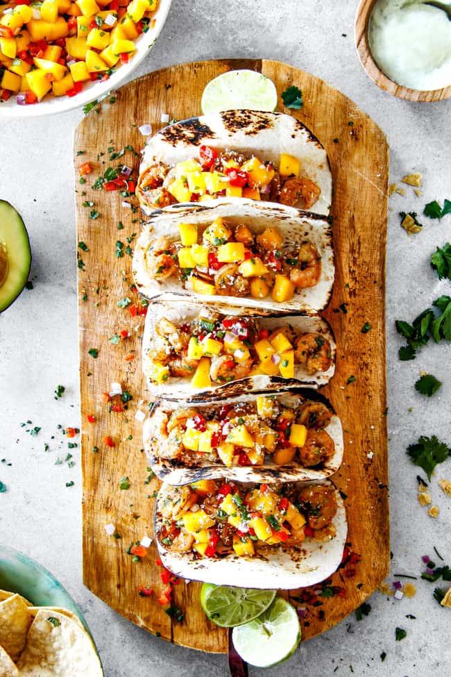 Cilantro Lime Shrimp Tacos EXPLODING with tangy, punchy flavor complimented by sweet refreshing Mango Salsa and silky Avocado Crema on your table in 35 minutes!ย You can prep the toppings ahead of time for a super quick and easy weeknight dinner that tastes better than any restaurant! easy Shrimp Tacos with Mango Salsa and Avocado lined in a row on a brown cutting board