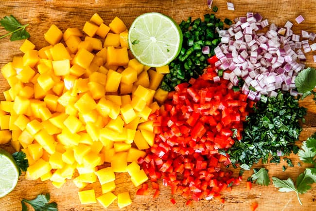 Cilantro Lime Shrimp Tacos EXPLODING with tangy, punchy flavor complimented by sweet refreshing Mango Salsa and silky Avocado Crema on your table in 35 minutes!ย You can prep the toppings ahead of time for a super quick and easy weeknight dinner that tastes better than any restaurant! showing how to make Shrimp Tacos by chopping mangos, red bell pepper, jalapeno, red onion, lime on a wood cutting board