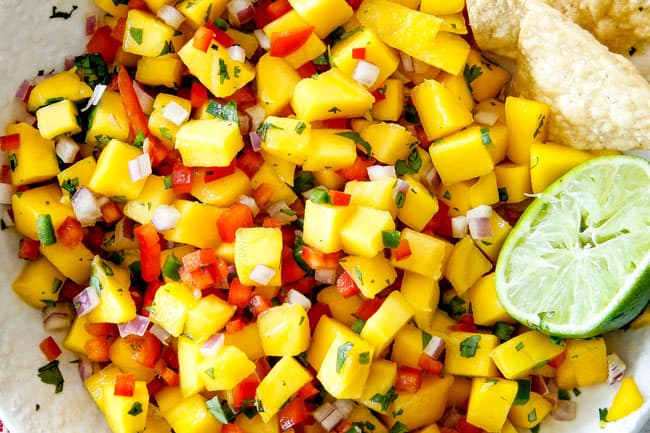 Cilantro Lime Shrimp Tacos EXPLODING with tangy, punchy flavor complimented by sweet refreshing Mango Salsa and silky Avocado Crema on your table in 35 minutes!ย You can prep the toppings ahead of time for a super quick and easy weeknight dinner that tastes better than any restaurant! showing how to make easy Shrimp Tacos by adding Mango Salsa to a white bowl