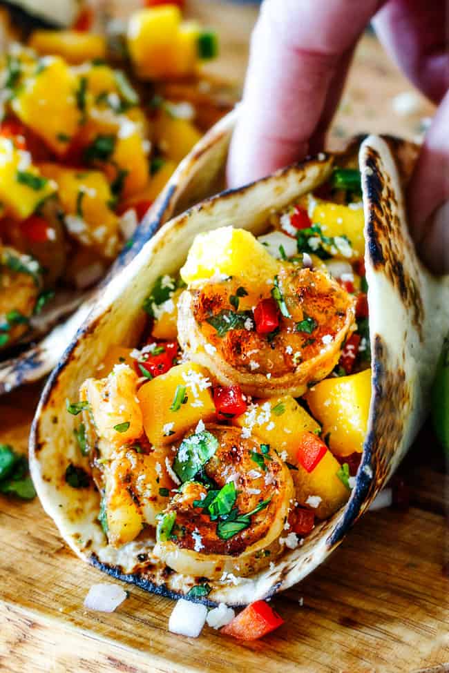 Cilantro Lime Shrimp Tacos EXPLODING with tangy, punchy flavor complimented by sweet refreshing Mango Salsa and silky Avocado Crema on your table in 35 minutes!ย You can prep the toppings ahead of time for a super quick and easy weeknight dinner that tastes better than any restaurant! up close of Shrimp Tacos with Mango Salsa and Avocado being grabbed by a hand to eat