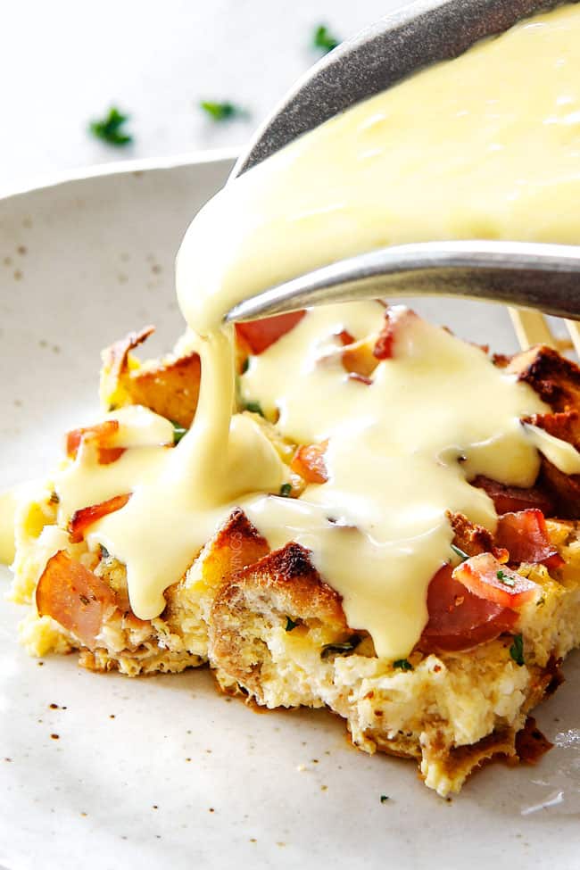 Eggs Benedict Casserole on a speckled plate with Hollandaise Sauce being poured from a silver pitcher over the slice