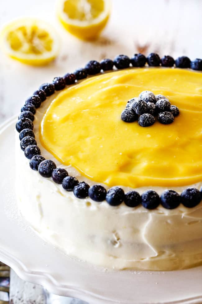 soft and tender Lemon Blueberry Cake with white lemon cream cheese frosting and blueberries around the edges on a white cake stand