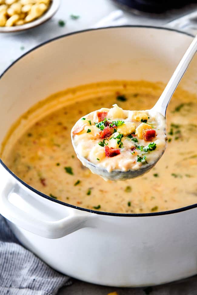 Better than any restaurant New England Clam Chowder that's SO EASY! Itโs loaded with tender clams, creamy potatoes and salty, smoky bacon in a rich flavorful chowder!ย You can use fresh OR canned clams and both versions will blow you away! side view of the best New England Clam Chowder recipe in the world in a white Dutch oven with potatoes, bacon, onions, celery and garlic with a bowl of oyster crackers