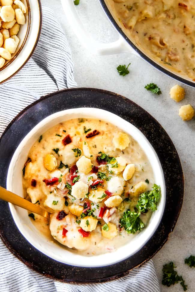 Better than any restaurant New England Clam Chowder that's SO EASY! Itโs loaded with tender clams, creamy potatoes and salty, smoky bacon in a rich flavorful chowder!ย You can use fresh OR canned clams and both versions will blow you away! top view of white bowl of easy New England Clam Chowder recipe with a spoon garnished with oyster crackers