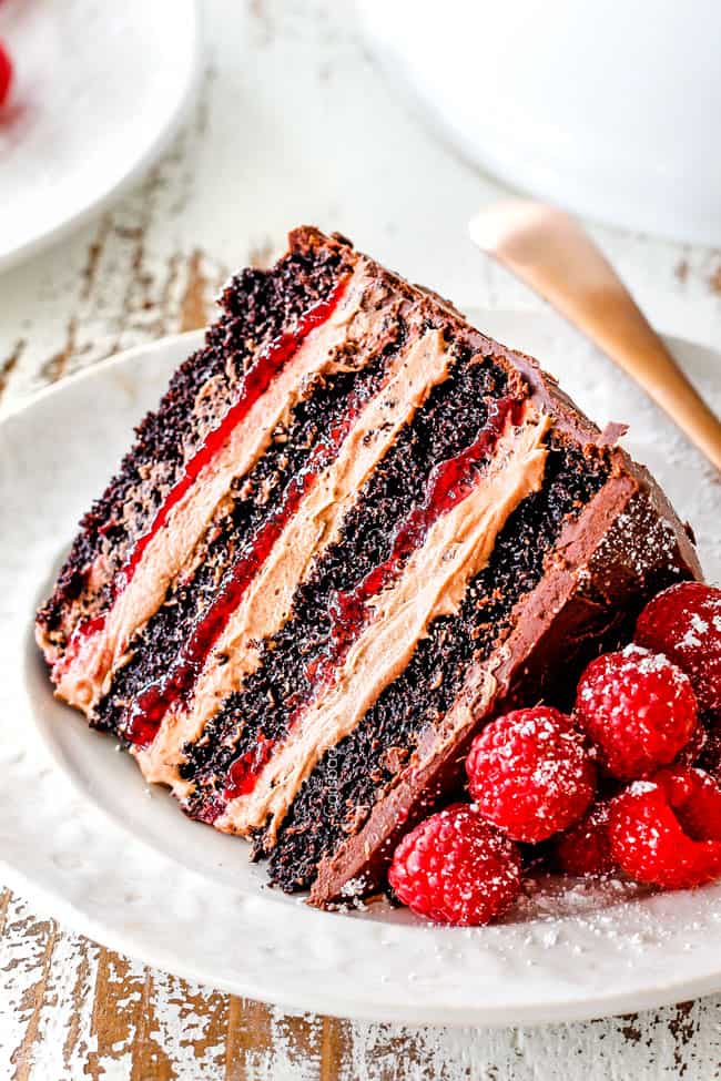 up close of a slice of Dark Chocolate Raspberry Cake on its side showing layers of dark chocolate cake, raspberry jam filling, chocolate ganache and chocolate mousse on a white plate with a gold fork 