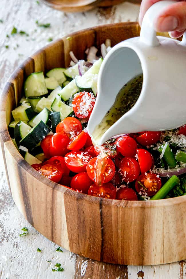 Pouring Italian Dressing from a white pitcher onto Tomato Cucumber Onion Salad in a wooden bowl