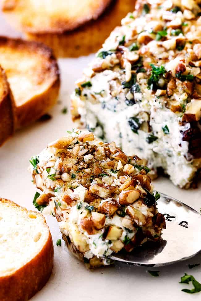 scooping part of cheese log with garlic, lemon zest, basil, chives, parsley and rolled in pecans with a knife