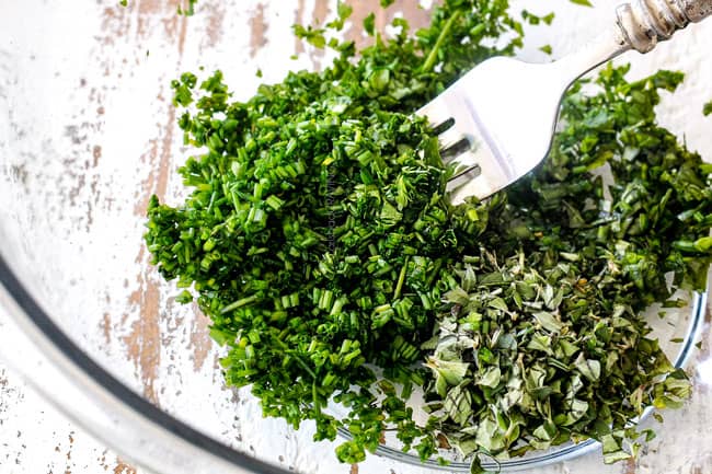 showing how to make cheese log by whisking together garlic, chives, basil, parsley, oregano in a glass bowl