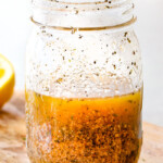 Homemade Italian Dressing in a glass mason jar on a wood cutting board with vinegar, oil, garlic, lemon juice, basil, parsley, oregano, salt and pepper