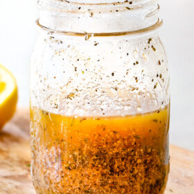 Homemade Italian Dressing in a glass mason jar on a wood cutting board with vinegar, oil, garlic, lemon juice, basil, parsley, oregano, salt and pepper