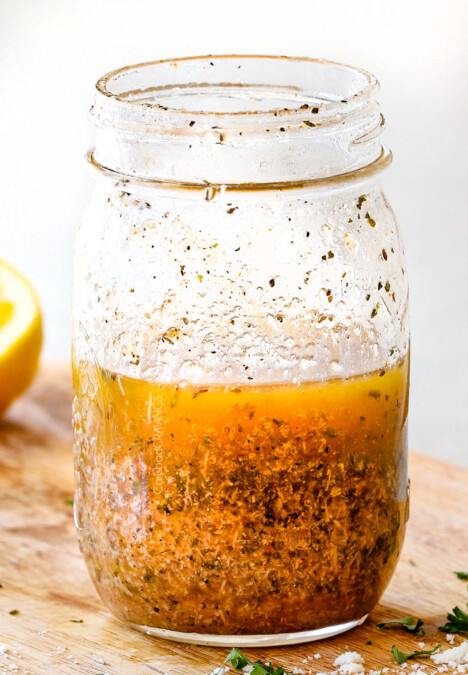Homemade Italian Dressing in a glass mason jar on a wood cutting board with vinegar, oil, garlic, lemon juice, basil, parsley, oregano, salt and pepper