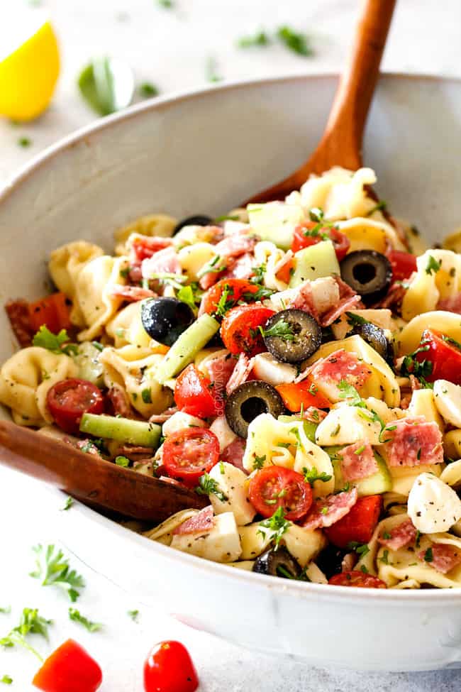 tossing together best Italian Pasta Salad with pepperoni, tomatoes, olives, cucumbers, mozzarella with wooden tongs