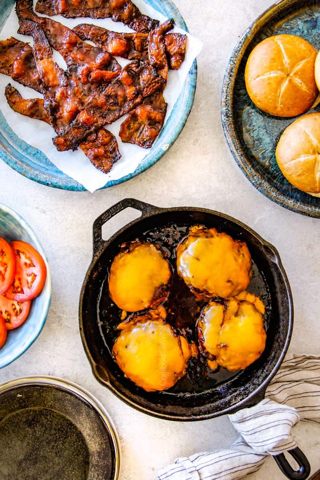 top view of four cheeseburgers in a skillet, bacon on a plate, tomatoes in a bowl and buns on aplate