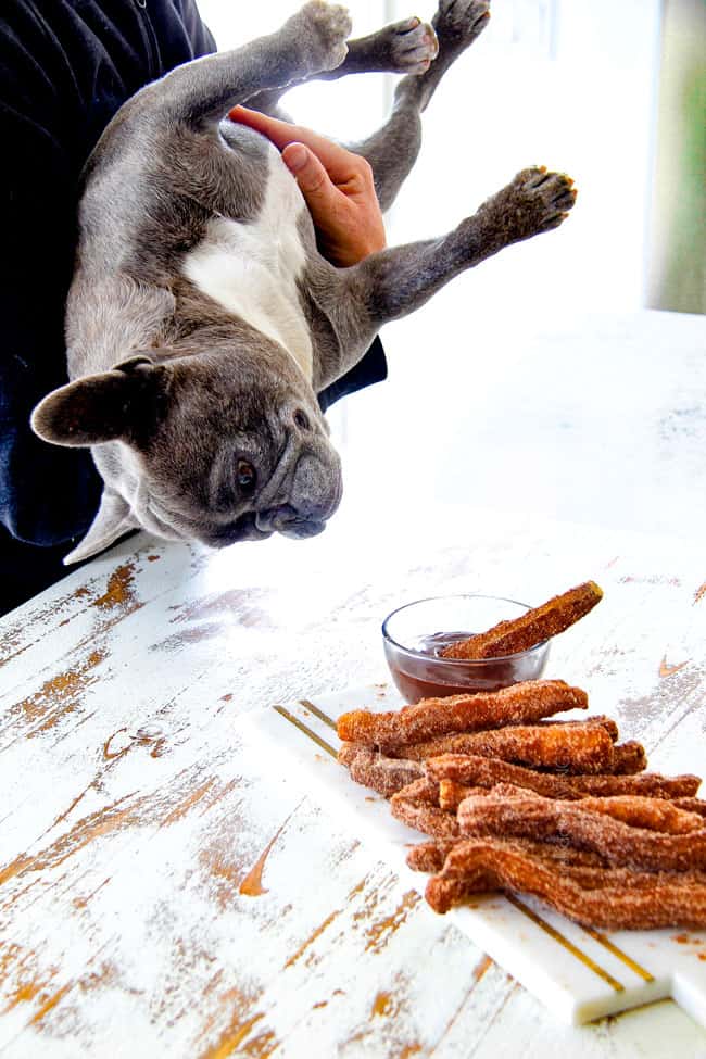 far away shot of one churro dipping in dark chocolate sauce in a glass bowl with stack of churros behind