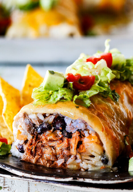 side view of wet burrito cut open with pulled pork, rice, black beans on a black plate garnished with lettuce, tomatoes, sour cream
