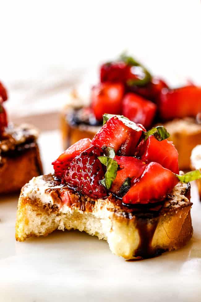 side view of strawberry goat cheese bruschetta on a white cutting board with a bite taken out