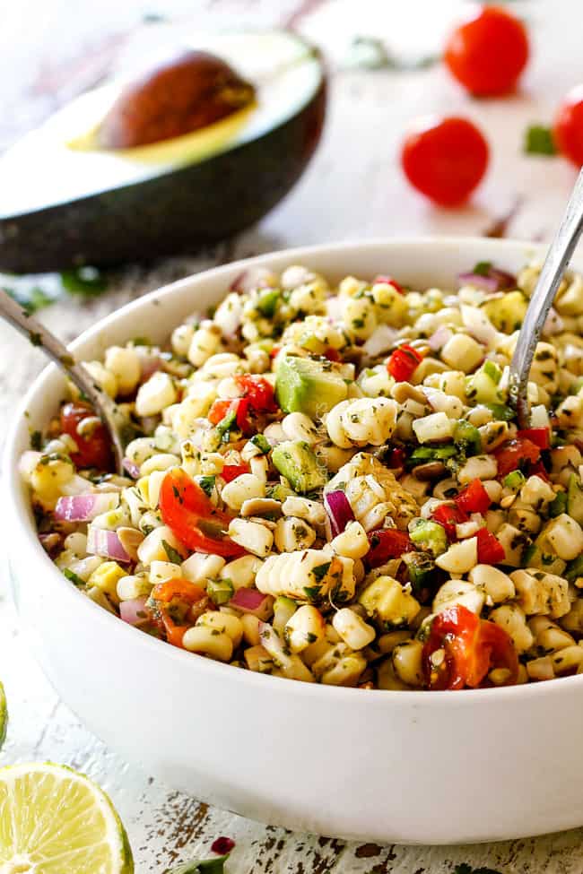 Tossing Fresh Corn Salad with avocado, red bell peppers, jalapeno, cilantro, sesame seeds mixed together in a white bowl