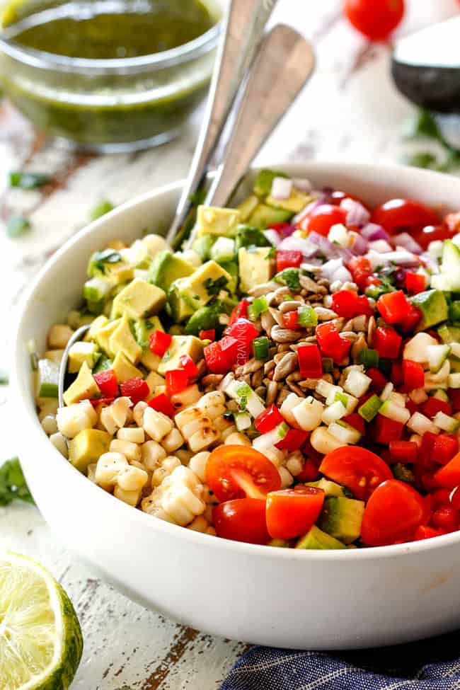 side view of corn salad ingredients in a white bowl with canned corn, avocados, tomatoes, jalapenos, cilantro, red onions, jalapeno, sesame seeds