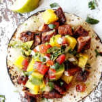 up close top view of al pastor pork on a corn tortilla with grilled pineapple, pico de gallo and avocado on a white board