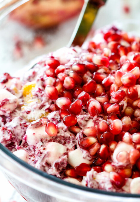up close of a spoon scooping classic ambrosia salad with pineapple, mandarin oranges, mini marshmallows, cranberries and pomegranate seeds in a glass bowl