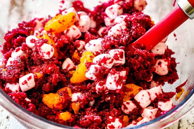 showing how to make ambrosia salad by stirring pineapple, mandarin oranges and cranberries together in a glass bowl