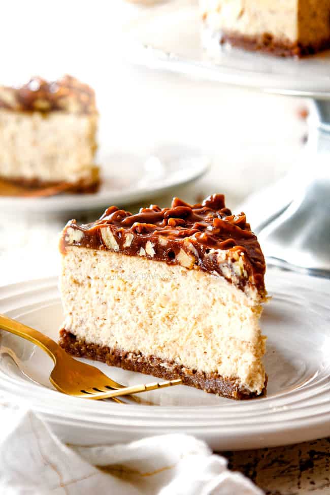 up close iew of side view of a slice of best pecan pie cheesecake on a white plate with a gold fork with the whole cheesecake on a pedestal