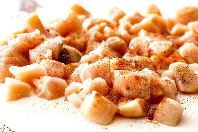 showing how to best chicken divan recipe by tossing chopped raw chicken with salt, pepper and paprika on a white cutting board