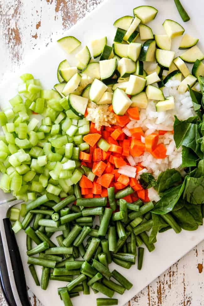 showing how to make hearty Minestrone Soup Recipe by chopping celery, carrots, onions, zucchini, green beans, garlic and onions on a white cutting board 