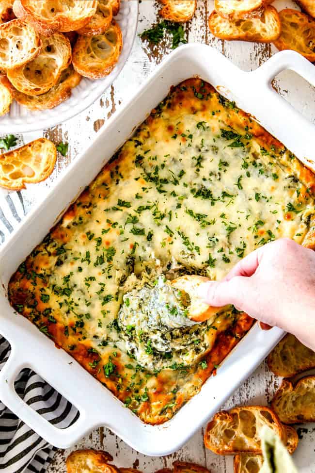 top view of a hand dipping a piece of bread into easy spinach artichoke dip