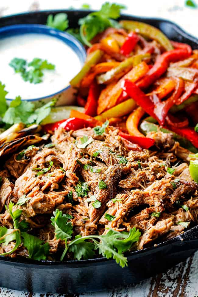up close side view of pork fajitas in a skillet with salsa verde pork, bell peppers, onions, limes, cilantro and tortillas